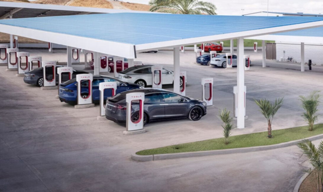 Useita Tesla-sähköautoja latauksessa sinisen katoksen alla modernilla Supercharger-asemalla.