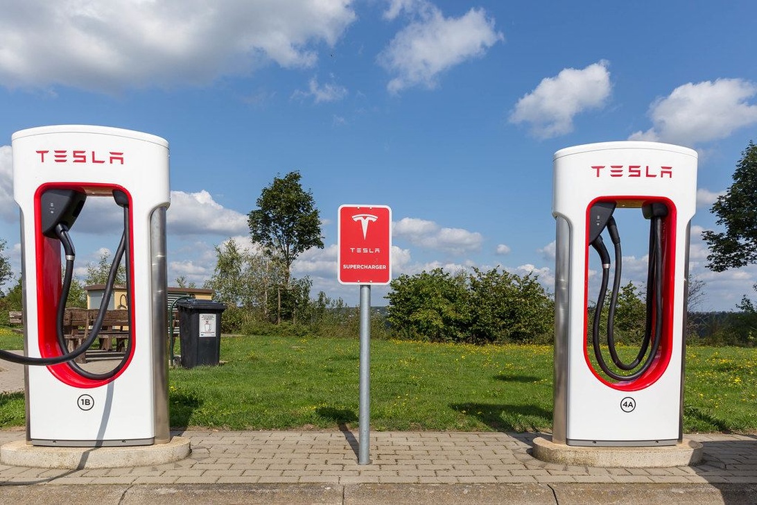 Kaksi valko-punaista Tesla Supercharger -latausyksikköä tiilipäällysteellä maaseutumaisemassa.